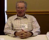 An older man wearing glasses sits at a table.