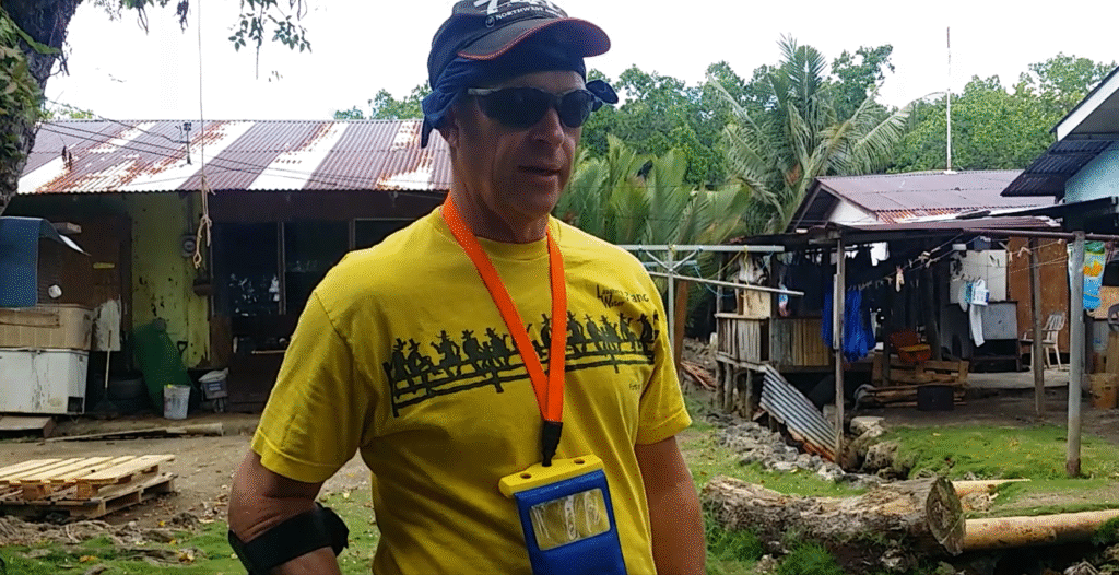 Man in yellow shirt outside wooden buildings.